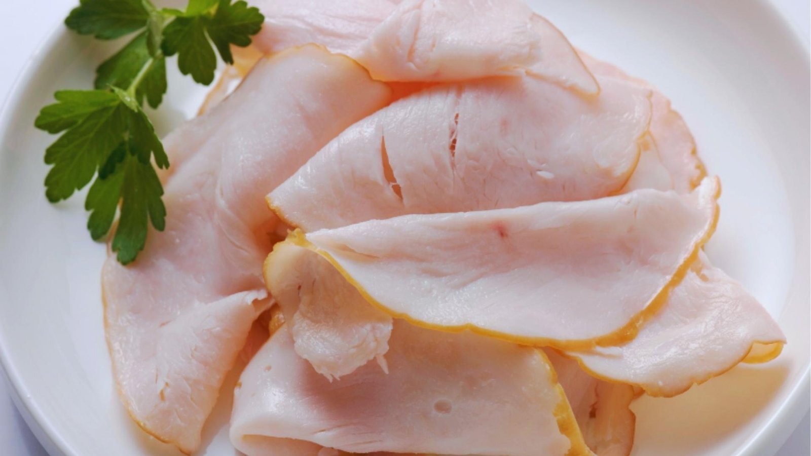 Thinly sliced deli turkey breast fanned out on white plate with fresh green parsley sprig for garnish.